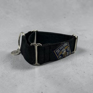 May include: A black dog collar with silver-colored hardware and a small circular tag. The collar features a rectangular patch with the text "THE POD SQUAD" in a navy blue and gold design. The collar is made of a dark fabric.