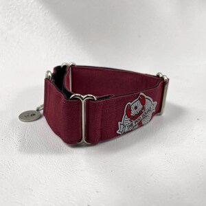 May include: A burgundy dog collar with a black interior and silver-toned hardware. The collar features a patch with the text "THE POD SQUAD" and a lobster graphic. A small, round charm hangs from the collar.