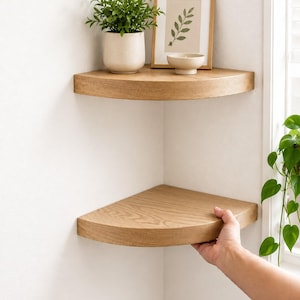 May include: Two wooden corner shelves mounted on a white wall. The top shelf holds a potted plant, a framed picture, and a small bowl. The bottom shelf is being held by a hand. The shelves are made of light-colored wood.