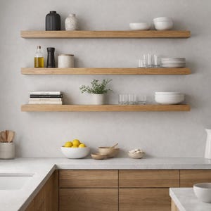 May include: Three horizontal light wood floating shelves mounted on a light gray wall above a kitchen counter. The shelves hold various kitchen items including ceramic bowls, glasses, a small potted plant, books, and containers for oil and spices.