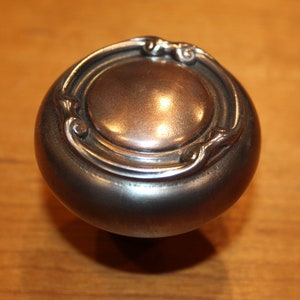 May include: A silver-toned door knob with a decorative, ornate design around the edge.