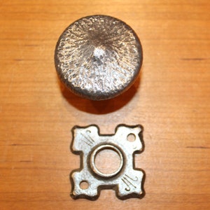 May include: A silver-colored, textured cabinet knob with a matching backplate. The knob is round with a slightly domed top. The backplate is square with decorative cutouts and a central hole for the knob's stem. Hardware for furniture.