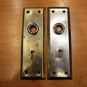 May include: Two antique door plates, one is brass and the other is silver. Both have a keyhole and a round knob hole.