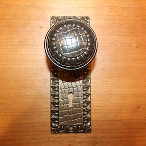 May include: A vintage door knob and backplate with a decorative, textured, alligator skin design. The knob is black and the backplate is silver.