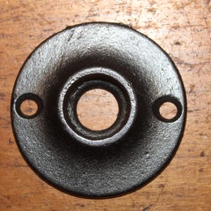 May include: A black, circular metal plate with a central hole and two smaller holes on either side. The plate has a textured surface and appears to be made of cast iron. It is likely a decorative or functional hardware piece.