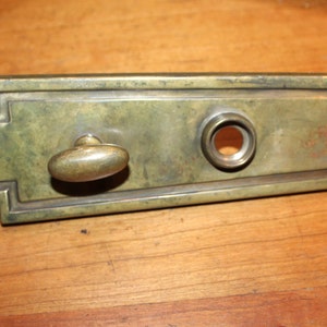 May include: A brass door plate with an Art Deco design. The plate has a round keyhole and a small oval knob.