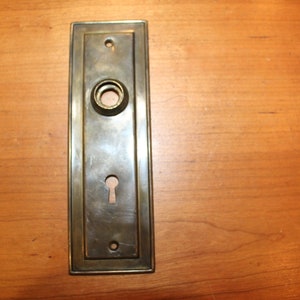 May include: A brass door plate with a keyhole and a round knob hole. The plate has a rectangular shape with a decorative border.