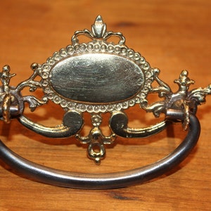 May include: Ornate brass drawer pull with a decorative oval center and a black metal loop handle.