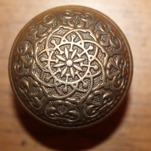 May include: A round, brass door knob with an intricate, ornate design. The knob has a raised, circular pattern in the center, surrounded by a decorative border.
