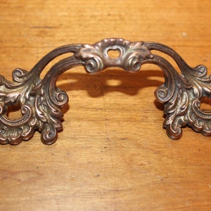 May include: Ornate brass drawer pull with a decorative, scroll-like design. The pull has a heart-shaped cutout in the center.
