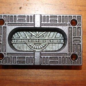 May include: A silver metal door plate with an ornate design. The plate has a rectangular shape with rounded corners and a raised oval in the center. The oval features a geometric pattern with a central sunburst design.