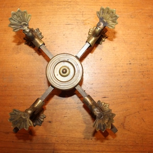 May include: An antique brass and metal decorative object. The piece has a central circular element with four arms extending outwards. Each arm terminates in a decorative, stylized floral design. The object is set against a wooden surface.