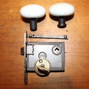 May include: Antique door hardware set featuring two white oval porcelain doorknobs with black stems, a black metal lock, and a brass keyhole. The lock has a decorative profile and a brass key. The set is displayed on a wooden surface.