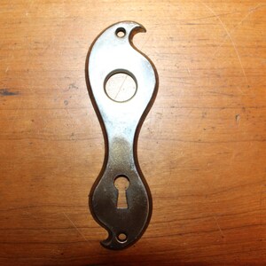 May include: A silver-colored metal door plate with a keyhole and a decorative cutout in the shape of a bird. The plate is on a wooden surface.