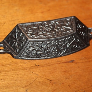 May include: A black, ornate drawer pull with a floral design. The cast iron handle has a curved shape with two mounting holes and is set against a wooden surface. The decorative hardware features detailed scrollwork.