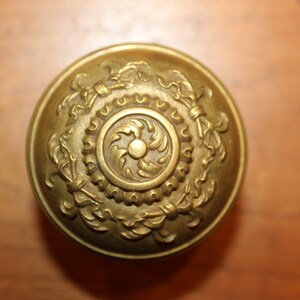 May include: A gold-toned metal door knob with an ornate design featuring a central circular motif and intricate floral patterns.