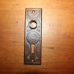May include: A vintage brass door plate with an ornate design. The plate has a keyhole and a round hole for a knob.