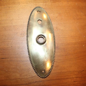 May include: A brass oval door plate with a beaded edge and three holes. The plate is slightly tarnished.