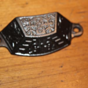 May include: A black, decorative drawer pull with a floral design in the center. The pull has a curved shape with two holes for mounting. The surface has a textured appearance, and the hardware is made of metal.