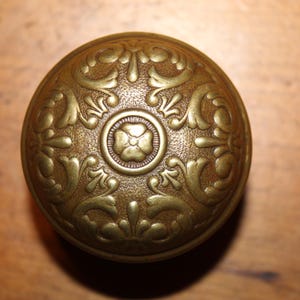 May include: Ornate, antique brass doorknob with a detailed floral design. The knob is round with a central flower motif and surrounding symmetrical leaf patterns. The brass has a warm, aged patina.