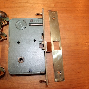 May include: A brass door knob set with a green metal door lock and a brass strike plate. The set includes two knobs, a lock, and a strike plate. The lock has a keyhole and a small metal lever. The strike plate has a rectangular opening for the latch.