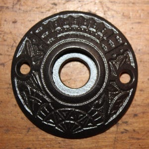 May include: A black metal door knob escutcheon with an ornate design. The escutcheon has a circular shape with a hole in the center for the knob.