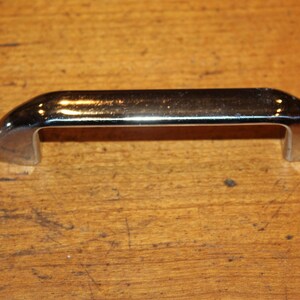 May include: A chrome-plated metal cabinet handle with a rounded rectangular shape.