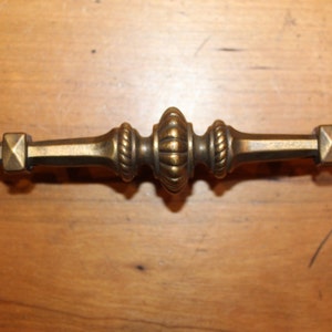 May include: A brass cabinet pull with a decorative design. The pull has a square shape on each end and a center section with a rope-like design.