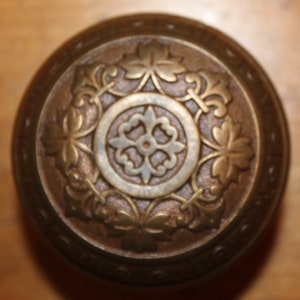 May include: A brass door knob with an intricate design featuring a central floral motif surrounded by stylized leaves. The knob has a circular shape and a slightly raised edge.