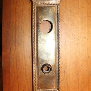 May include: A gold-toned metal door plate with an ornate design and two keyholes. The plate has a rectangular shape with a decorative top and bottom.