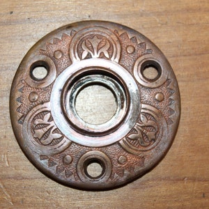 May include: A round, ornate, copper-colored door knob with a decorative design. The knob has a circular opening in the center and four smaller holes around the edge.