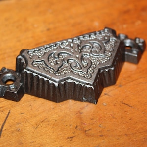 May include: A black metal door handle with an ornate design. The handle has a decorative, raised pattern on the top and a smooth, rounded surface on the bottom.