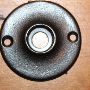May include: A black metal door knob escutcheon with a round shape and a smooth, slightly textured surface. The escutcheon has a central hole for the knob and two smaller holes on the sides.
