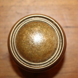May include: Close-up of a round, antique brass cabinet knob. The knob has a domed center and concentric rings around the edge. The finish is aged with a slightly worn appearance. The background is a wooden surface.