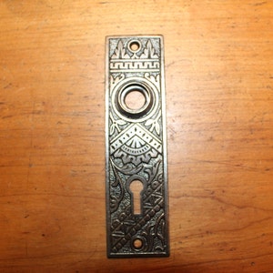 May include: A vintage, ornate, silver-toned door plate with a keyhole and a round knob. The plate has a decorative design with intricate details.