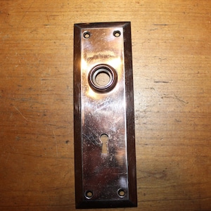 May include: A vintage brass door plate with a keyhole and a round knob. The plate is surrounded by a dark brown wooden frame.