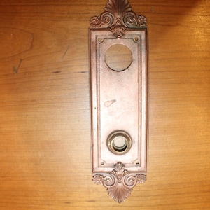 May include: A vintage, ornate, copper-colored door plate with a circular cutout and a keyhole. The plate has a decorative, scroll-like design around the edges.