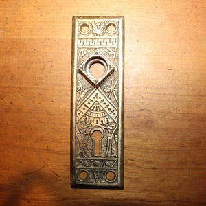 May include: An antique brass door plate with intricate floral and geometric patterns. The rectangular plate features a keyhole, two circular holes, and a diamond-shaped opening. The design includes detailed engravings and a vintage aesthetic.