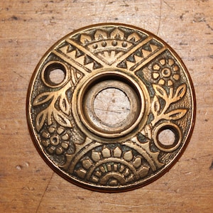 May include: Ornate brass door plate with floral and geometric patterns. The circular plate features a central hole and two smaller holes, with detailed relief designs of flowers and leaves. The antique-style plate is set against a wooden surface.
