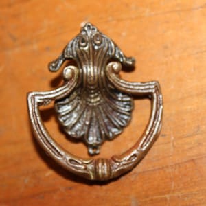 May include: A vintage, ornate, metal drawer pull with a shell-shaped design. The pull is a dark bronze color and has a decorative, floral-like top.