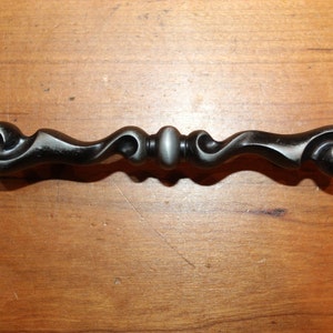 May include: A black metal cabinet pull with a decorative, ornate design. The pull has a curved shape with a central knob.