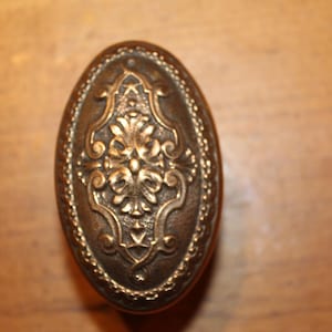 May include: Oval-shaped antique brass door knob with ornate floral and scroll detailing. The knob has a raised central design and a beaded border. A classic hardware piece for vintage or traditional decor.
