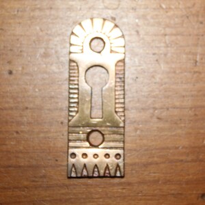 May include: A brass-colored keyhole cover with an arched top and decorative details. The cover has a keyhole in the center and two small holes, one above and one below the keyhole. The bottom edge features a row of triangles.