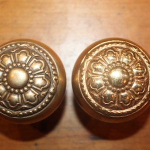 May include: Two antique brass door knobs with a floral design. The knobs are round and have a raised center with a smaller circle in the middle. The knobs are surrounded by a decorative border.