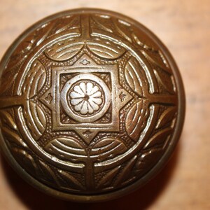 May include: A round, brass door knob with an intricate, geometric design. The knob has a raised, circular pattern in the center.