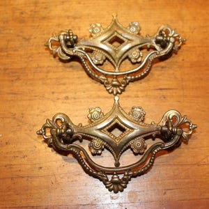 May include: Two antique brass drawer pulls with ornate floral and diamond-shaped designs. The pulls have a curved handle and are set against a wooden surface. These decorative hardware pieces add a vintage touch.