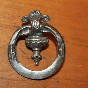 May include: A silver-toned metal ring pull with a decorative floral design. The ring pull has a round shape with a raised center piece that resembles a vase.