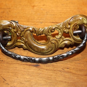 May include: Ornate antique drawer pull with a brass-colored decorative backplate and a dark gray handle. The backplate features intricate scrollwork and a curved design. The handle is a curved bar with a textured surface.
