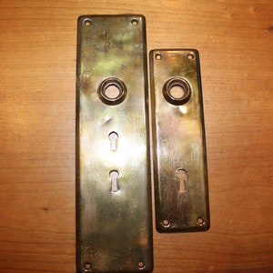 May include: Two antique brass door plates. The larger plate has a keyhole and two screw holes at the top and bottom. The smaller plate has a keyhole and two screw holes. Both plates have a circular opening.
