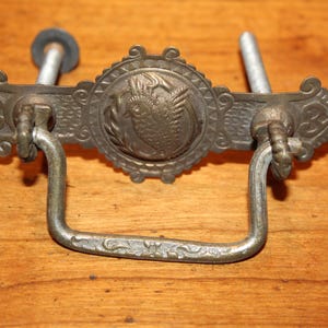 May include: Antique metal drawer pull with an ornate design. The center features a circular emblem with a bird motif. The pull has a curved handle and two mounting posts. The metal has a tarnished, aged appearance.